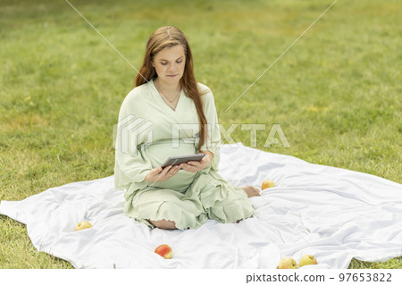 Young pretty pregnant woman holding, reading something on tablet,gadget,resting in park,sitting on cover,blanket in park,meadow. Healthy walk outdoors. Preparation for childbirth. Horizontal Young pretty pregnant woman holding, reading something on tablet,gadget,resting in park,sitting on cover,blanket in park,meadow. Healthy walk outdoors. Preparation for childbirth. Horizontal 97653822