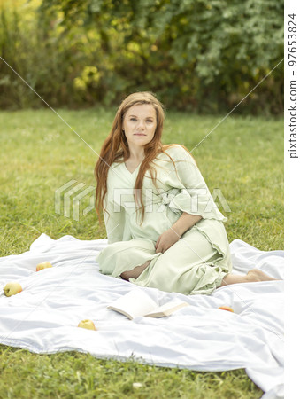 Young pretty pregnant woman resting in park, lying on cover,blanket in park,meadow, enjoying pregnancy, apples and book are beside. Healthy walk outdoors. Preparation for childbirth. Vertical 97653824
