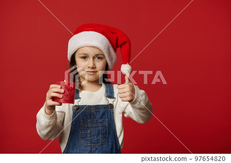 Beautiful child girl in white pullover, blue denim overalls and Santa hat, holding a Christmas candle and showing thumb up looking at camera, isolated over red background with copy advertising space Beautiful child girl in white pullover, blue denim overalls and Santa hat, holding a Christmas candle and showing thumb up looking at camera, isolated over red background with copy advertising space 97654820