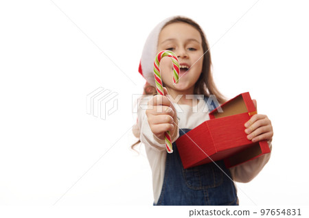 Focus on red green striped lillipop in the hands of a blurred overjoyed happy smiling child girl in Santa hat, with gift box, holding out a sweet candy cane to camera, isolated over white background Focus on red green striped lillipop in the hands of a blurred overjoyed happy smiling child girl in Santa hat, with gift box, holding out a sweet candy cane to camera, isolated over white background 97654831