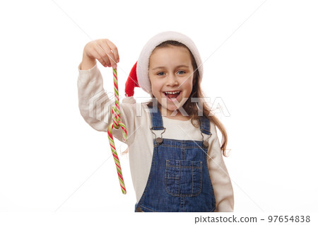 Caucasian smiling cheerful little girl wearing blue denim overalls and red Santa hat, playing with striped red and green candy canes lollipops on white background. Christmas New Year winter Holidays Caucasian smiling cheerful little girl wearing blue denim overalls and red Santa hat, playing with striped red and green candy canes lollipops on white background. Christmas New Year winter Holidays 97654838