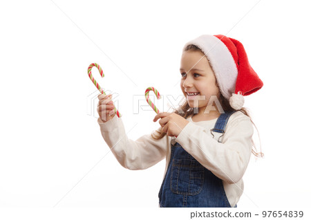 Caucasian 5-6 years child, cute baby girl in Santa hat, pointing with green red striped candy canes at copy advertising space for Christmas, on white background, expressing happiness and cheerfulness 97654839