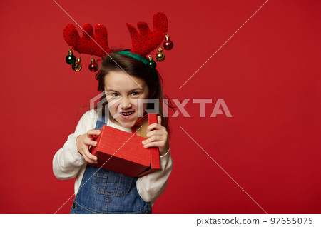 Mischievous funny little child girl in deer antler hoop, white pullover and blue denim overalls, smiling looking at camera, unpacking gift box with surprise for Christmas, on red background. Ad space Mischievous funny little child girl in deer antler hoop, white pullover and blue denim overalls, smiling looking at camera, unpacking gift box with surprise for Christmas, on red background. Ad space 97655075