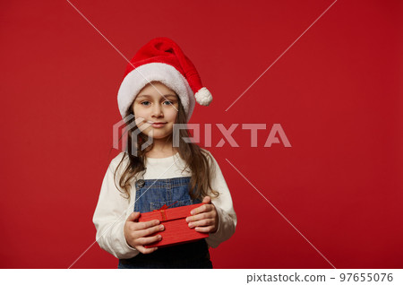 Lovely Caucasian little child girl in Santa hat, white pullover and blue denim overalls, smiling looking at camera, posing with cute gift box with surprise for Christmas, on red background. Ad space Lovely Caucasian little child girl in Santa hat, white pullover and blue denim overalls, smiling looking at camera, posing with cute gift box with surprise for Christmas, on red background. Ad space 97655076