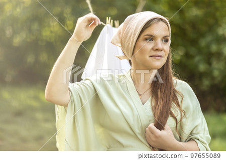 Portrait of young pretty white woman hanging up laundry on clothes line outside, drying bed linens. Safe laundry detergents, organic washing gel, fabric softener concept. Skin, health care. Horizontal Portrait of young pretty white woman hanging up laundry on clothes line outside, drying bed linens. Safe laundry detergents, organic washing gel, fabric softener concept. Skin, health care. Horizontal 97655089