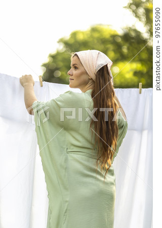 Young pretty white woman hanging up laundry on clothes line outside, drying bed linens, bedding. Safe laundry detergents, organic washing gel, fabric softener concept. Skin, health care. Vertical Young pretty white woman hanging up laundry on clothes line outside, drying bed linens, bedding. Safe laundry detergents, organic washing gel, fabric softener concept. Skin, health care. Vertical 97655090