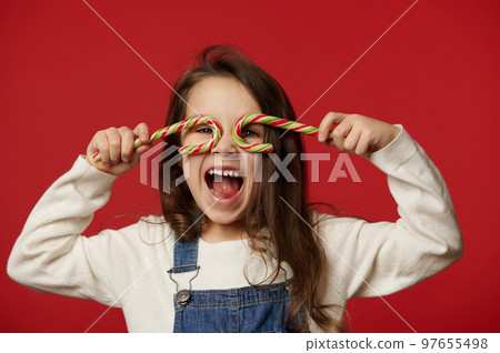 Funny amazed cheery little child girl, looking through striped lollipops like eyeglasses, laughing looking at camera, isolated over red background with free advertising space for text. Merry Christmas 97655498