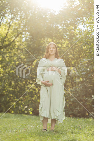Young pregnant woman puts hands on big belly, green trees,sky on background. Brown haired female wears dress. Magic happy pregnancy. Child delivery preparation,emotional connection with baby. Vertical 97655844
