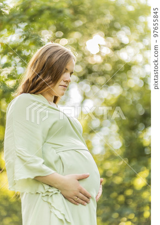 Young pregnant woman holds big belly, green trees,sky on background. Brown long haired female wear dress. Magic happy pregnancy. Child delivery preparations,emotional connection with baby. Vertical 97655845