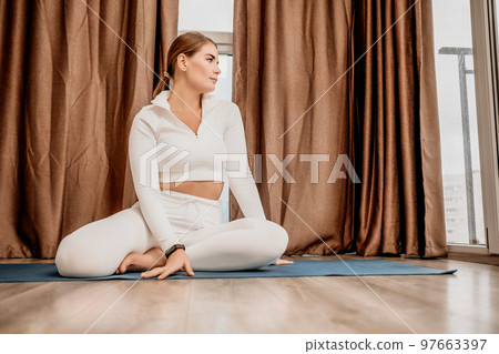 Well looking middle aged woman with long hair, fitness instructor in white sportswear, doing stretching and pilates on yoga mat at home. Healthy lifestyle, harmony, fitness yoga and meditation. 97663397