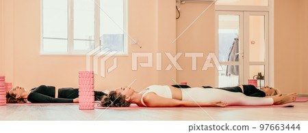 Middle aged well looking women, performing fascia exercises on the floor using a massage foam roller - tool to relieve tension in the back and relieve muscle pain. Female fitness yoga routine concept Middle aged well looking women, performing fascia exercises on the floor using a massage foam roller - tool to relieve tension in the back and relieve muscle pain. Female fitness yoga routine concept 97663444