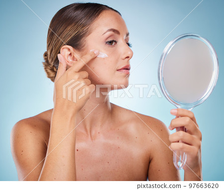Woman, face and cream with hand mirror for beauty wellness, skincare dermatology and cosmetics moisturizer in blue background studio. Model, apply sunscreen and makeup facial product for self care 97663620