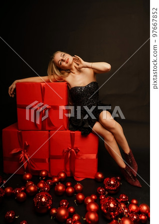 Woman gifts christmas. Beautiful sexy young blonde in a black dress sits on three big red gifts on a black background. Christmas photo, gifts Woman gifts christmas. Beautiful sexy young blonde in a black dress sits on three big red gifts on a black background. Christmas photo, gifts 97667852