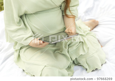 Cropped pregnant woman holding, reading something on tablet, gadget, resting in park,sitting on blanket in park, meadow. Healthy walk outdoors. Preparation for childbirth. Horizontal Cropped pregnant woman holding, reading something on tablet, gadget, resting in park,sitting on blanket in park, meadow. Healthy walk outdoors. Preparation for childbirth. Horizontal 97668269