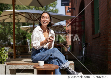 Stylish young asian woman sitting with cup of coffee in an outdoor cafe, smiling and looking happy, looking around Stylish young asian woman sitting with cup of coffee in an outdoor cafe, smiling and looking happy, looking around 97669659