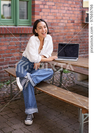 Vertical shot of good-looking asian woman with laptop, sitting in outdoor cafe, drinking coffee and smiling, laughing carefree 97669766