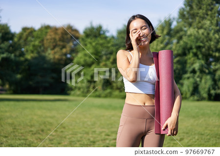 Portrait of asian girl smiles and laughs, stands with sports equipment, rubber mat, wears uniform for workout and exercises in park Portrait of asian girl smiles and laughs, stands with sports equipment, rubber mat, wears uniform for workout and exercises in park 97669767