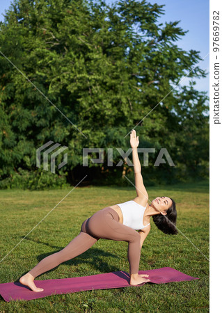 Vertical shot of young korean woman doing yoga training on rubber mat, making asana exercises on fresh air in park 97669782