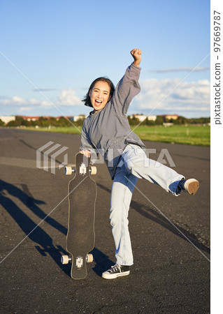 Vertical shot of asian girl feeling excited, skating on longboard, jumping and posing with skateboard, standing with cruiser on empty road, having fun outdoors 97669787