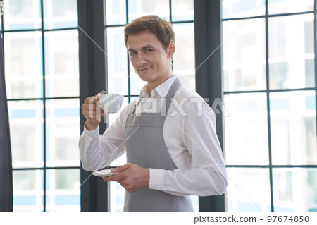 Portrait of a successful young man working as a professional barista, smiling at the camera and raising a cup with a coffee drink. Positive male waiter. High quality photo Portrait of a successful young man working as a professional barista, smiling at the camera and raising a cup with a coffee drink. Positive male waiter. High quality photo 97674850