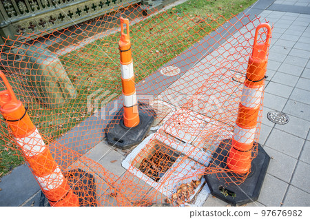 Open cover on a sidewalk access panel with hazard cones and fencing. Open cover on a sidewalk access panel with hazard cones and fencing. 97676982