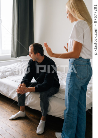 Vertical shot of despaired young husband sitting on bed looking away ignoring angry blonde wife arguing blaming upset man of problems at home. Jealous blonde woman shouting at sad boyfriend. 97677291