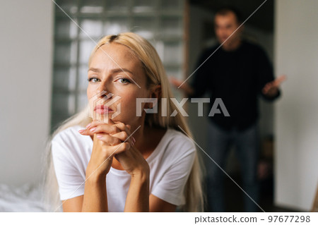 Portrait of tired young woman sadness looking at camera sitting on bed on background of aggressive husband shouting on girlfriend at home. Concept of family scandal, crisis, domestic violence, abuse. 97677298