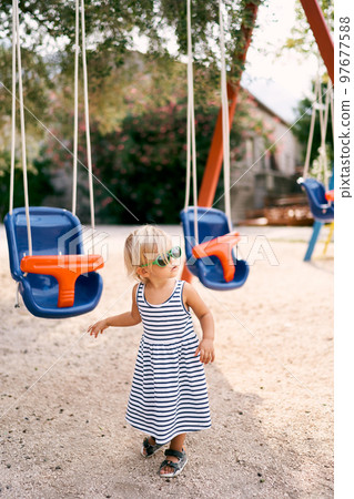Little girl in sunglasses walks past colorful swings. High quality photo Little girl in sunglasses walks past colorful swings. High quality photo 97677588