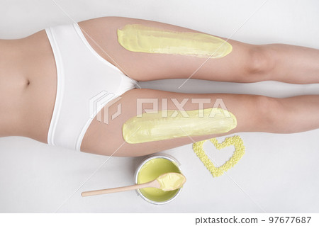 Top view of green hot wax on legs of slim woman while she lying down on couch. Depilation process in professional beauty salon. Healthy slim female body in white panties. Part of photo series. Top view of green hot wax on legs of slim woman while she lying down on couch. Depilation process in professional beauty salon. Healthy slim female body in white panties. Part of photo series. 97677687