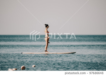 Sea woman sup. Silhouette of happy middle aged tanned woman in rainbow bikini, surfing on SUP board, confident paddling through water surface. Idyllic sunset. Active lifestyle at sea or river. 97686648