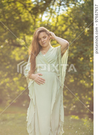 Young pregnant woman stands in park,looks at belly, green trees on background. Brown haired female wears dress.Magic happy pregnancy. Childbirth preparation, emotional connection with baby.Vertical 97687657