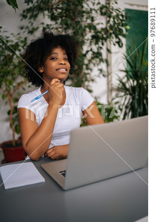 Vertical portrait of confident curly African female student woman holding pen in hand, smiling looking at camera sitting at table in light home office room with modern biophilic interior design. 97689111