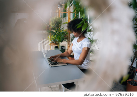 Side view of African-American female freelancer designer creating new fresh ideas, working on laptop computer in home office room with biophilic interior design, on background of green plants. 97689116