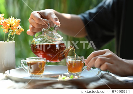 Cropped view of young man pouring traditional tea from transparent teapot into a glass cup 97690954