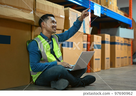 Handsome male worker sitting in retail warehouse full of cardboard boxes and using laptop computer 97690962