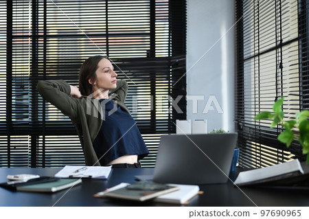 Thoughtful businesswoman relaxing on office chair with hands behind her head and looking through window Thoughtful businesswoman relaxing on office chair with hands behind her head and looking through window 97690965