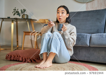 Portrait of girl watching television at home, sits on floor near sofa, holds remote and changes channels Portrait of girl watching television at home, sits on floor near sofa, holds remote and changes channels 97691944