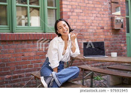 Happy korean girl with laptop, sitting in cafe, laughing and pointing at copy space on brick wall, showing advertisement Happy korean girl with laptop, sitting in cafe, laughing and pointing at copy space on brick wall, showing advertisement 97691945