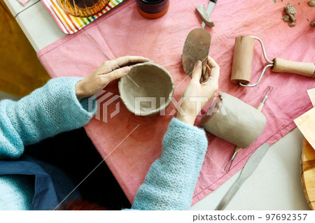 Women's hands knead the clay and sculpt a cup or bowl from it. The process of manufacturing a ceramic product,top view Women's hands knead the clay and sculpt a cup or bowl from it. The process of manufacturing a ceramic product,top view 97692357