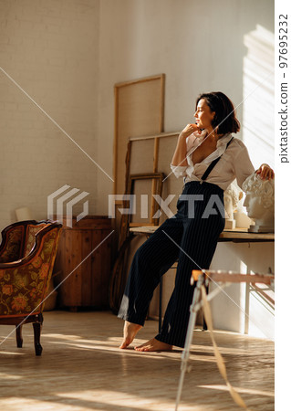 a brunette in a white shirt and trousers with suspenders in the artist's studio. 97695232