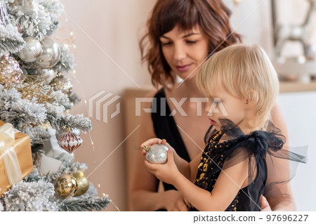 A mother with a 2 years old daughter decorates the Christmas tree. Both are dressed in black dresses, the daughter hangs a ball on the Christmas tree. 97696227