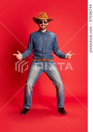 Excited young man in cowboy style outfit having fun, posing isolated over red background. Model in cowboy hat and denim clothes. Fashion, emotions concept 97696744