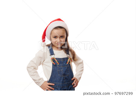 Adorable Caucasian little child girl wearing Santa hat, posing with hands on waist, with Christmas striped candy canes on the pocket of his denim overalls, looking at camera, against white background Adorable Caucasian little child girl wearing Santa hat, posing with hands on waist, with Christmas striped candy canes on the pocket of his denim overalls, looking at camera, against white background 97697299