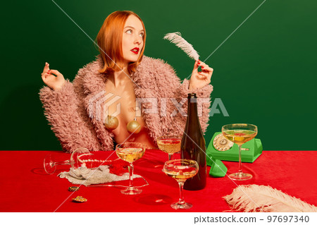 Portrait of beautiful redhead girl in pink fur coat sitting at the table with champagne over green background 97697340