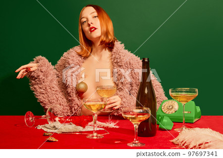 Portrait of beautiful redhead girl in pink fur coat sitting at the table with champagne over green background. New year celebration 97697341