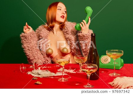 Portrait of beautiful redhead girl in pink fur coat sitting at the table with champagne over green background. Merry Christmas. 97697342