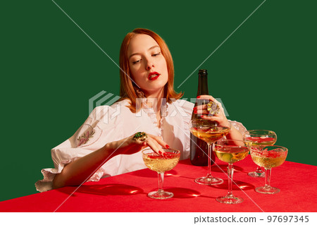Young redhead girl in festive white dress sitting at the table with champagne over green background. Luxury party Young redhead girl in festive white dress sitting at the table with champagne over green background. Luxury party 97697345