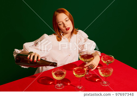 Young redhead girl in festive white dress sitting at the table and pouring champagne into glass over green background. 97697348