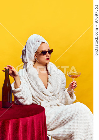Young woman in bathrobe sitting and drinking champagne over yellow background. Feeling relaxed 97697353