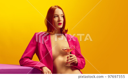 Drink aesthetics. Young redhead woman in pink jacket holding glass with champagne over yellow background Drink aesthetics. Young redhead woman in pink jacket holding glass with champagne over yellow background 97697355